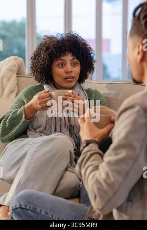 African American couple holding ceramic mugs and chatting on sofa in living room Stock Photo