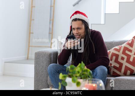 African American man wearing Santa hat holding smartphone near mouth on sofa by candle, copy space Stock Photo