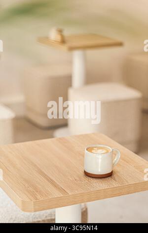 A ceramic cup of cappuccino with elegant cream latte art sits on a light wooden table in a minimalist coffee shop setting. Stock Photo