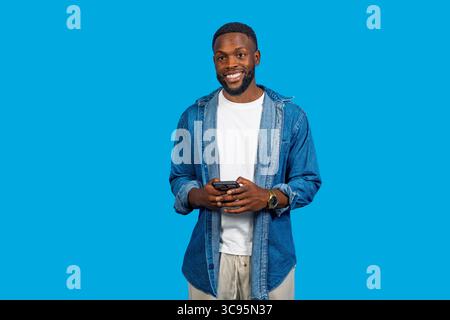 Young man using mobile phone, smiling and looking away on blue studio background Stock Photo