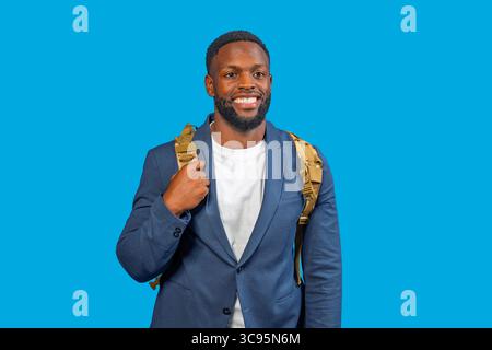 Portrait of a cheerful businessman carrying a backpack, exuding confidence and professionalism against a vibrant blue backdrop Stock Photo