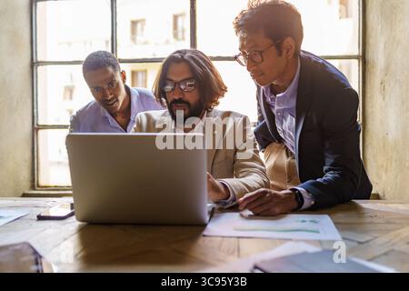 A focused multi-ethnic team of three male professionals collaborates on a business project, looking at a laptop screen during a meeting in a modern lo Stock Photo