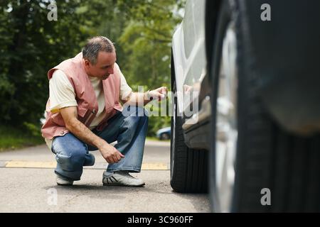 Middle aged Caucasian man crouching beside car inspecting front tire during family road trip on rural road, focusing on checking for damage or low pressure, trees visible in background Stock Photo
