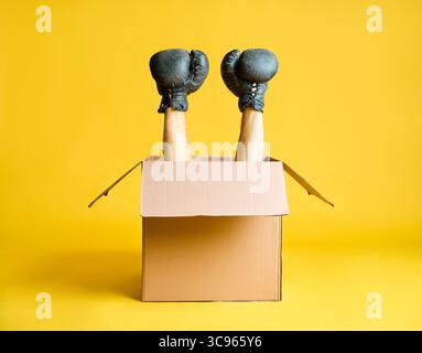 Pair of hands in boxing gloves raised up from open cardboard box on yellow background. Stock Photo