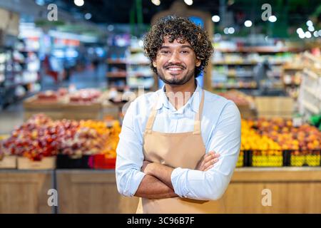 A smiling male grocery store worker stands with arms crossed in front of a fruit display, exuding confidence and friendliness. Stock Photo