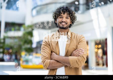 A smiling man with curly hair poses, arms crossed, inside a modern, blurred background. Stock Photo