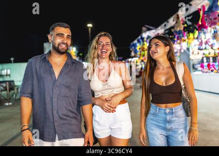 Three friends are enjoying their time together, strolling through a vibrant amusement park at night Stock Photo