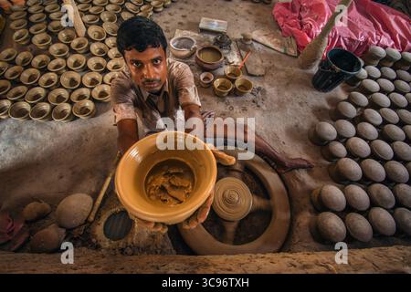 Bogura, Bangladesh - 20 May 2017: View of a potter, his hands showcasing a freshly molded clay pot, surrounded by rows of earthenware against a backdrop of raw materials. Stock Photo