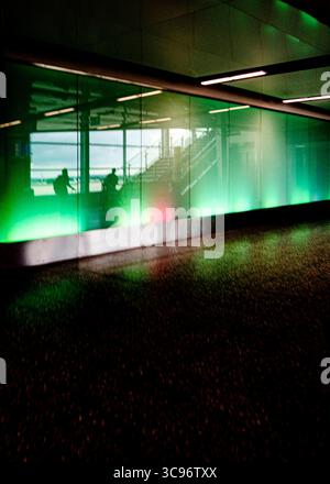 Two silhouetted figures are seen through a glass wall, possibly in a transit area. The wall is illuminated with green and red lights. Stock Photo