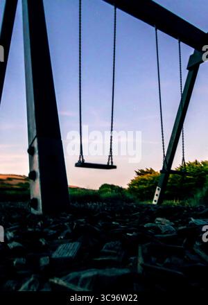 An empty swing set stands silhouetted against a dusky sky, with the ground covered in dark mulch. Stock Photo