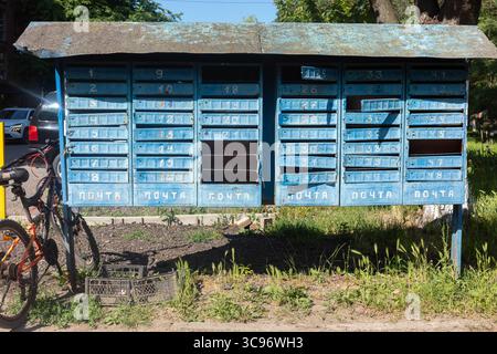 An old, blue, weathered post office box with multiple compartments, with a bicycle parked nearby, set against a backdrop of greenery and a sunny day. Stock Photo