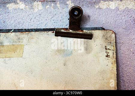 Macro view of a rusty metal clip attached to an aged and stained sketch board against a worn and textured purple wall. Stock Photo