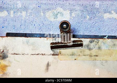 Macro view of a rusty metal clip attached to an aged and stained sketch board against a worn and textured purple wall. Stock Photo
