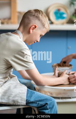 Blond boy learning pottery, close-up of hands shaping clay on a wheel, creative kids activity in cozy pottery studio, teamwork and manual craft skills Stock Photo