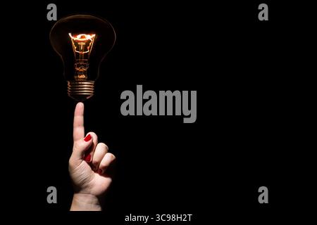 Idea and success concept. Glowing light bulb without wires on female hand with trigger finger and red nails on black background. Copy space Stock Photo