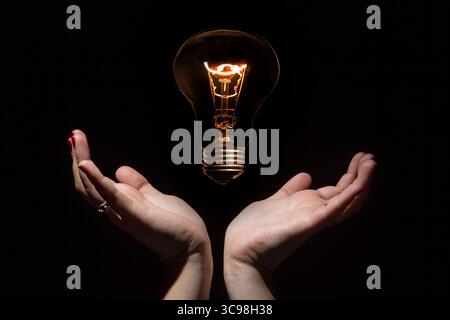 Idea and success concept. Glowing light bulb without wires and opened female hands on black background Stock Photo
