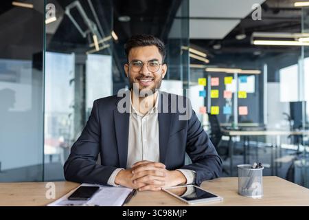 A smiling businessman sits at a desk in a modern office. He wears glasses and a suit, appearing confident and professional. Stock Photo