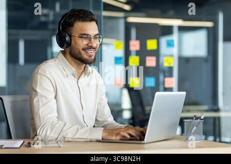 A smiling man wearing headphones uses a laptop in a modern office setting. He has glasses and is working on a project. Stock Photo