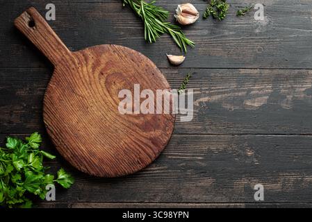 Empty wooden round board with herbs and garlic on dark rustic background, top view, copy space. Wooden pizza platter. Food background, design template Stock Photo