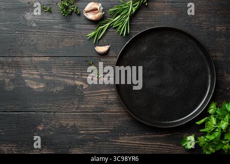 Empty black ceramic plate with herbs and garlic on dark wooden background, top view, copy space. Serving platter mockup. Food background, design templ Stock Photo