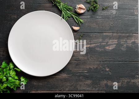 Empty white ceramic plate with herbs and garlic on dark wooden background, top view, copy space. Serving platter mockup. Food background, design templ Stock Photo
