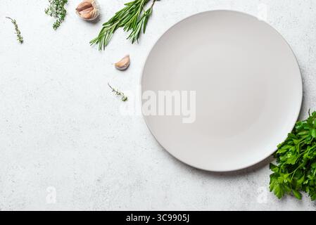 Empty white ceramic plate with herbs and garlic on white concrete background, top view, copy space. Serving platter mockup. Food background, design te Stock Photo
