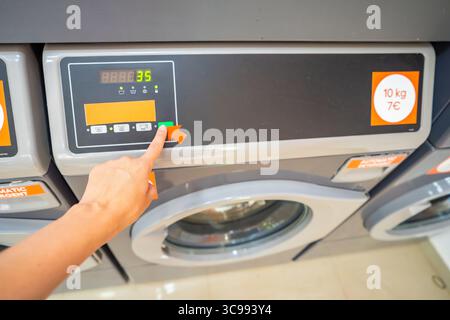 Close-up of female hand selecting washing program on modern washing machine. Choosing laundry mode in public self-service facility. Stock Photo