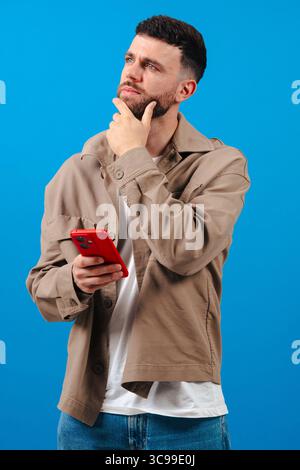 Pensive young man with smartphone over blue backdrop, planning next message idea Stock Photo