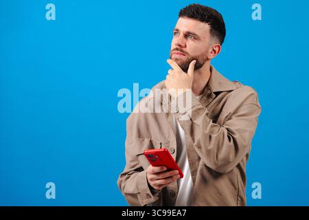 Pensive young man with smartphone over blue backdrop, planning next message idea Stock Photo