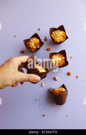A top-down view of a hand reaching for a delicious freshly baked muffin or cupcake in a brown paper liner, with other muffins and nuts scattered Stock Photo