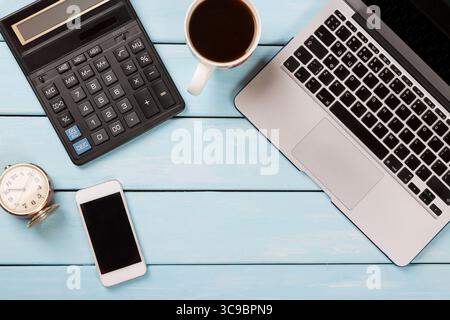 Business concept. Working desk with Laptop, calculator, modern phone, coffee nd vintage alarm clock on blue wooden table Stock Photo