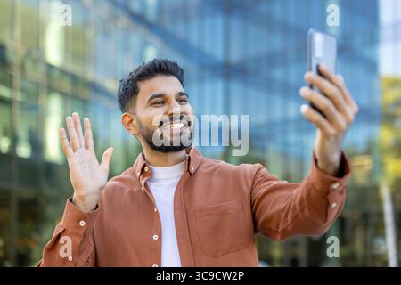 A smiling man waves while holding a phone, making a video call in front of a modern building. Stock Photo