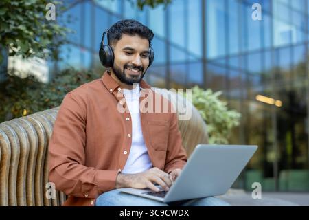 An Indian man wearing headphones smiles while using a laptop outdoors on a bench, surrounded by greenery. Stock Photo
