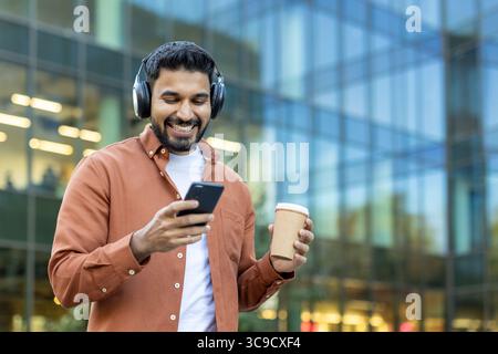 A smiling man enjoys music and coffee while using his phone in front of a modern building. Stock Photo