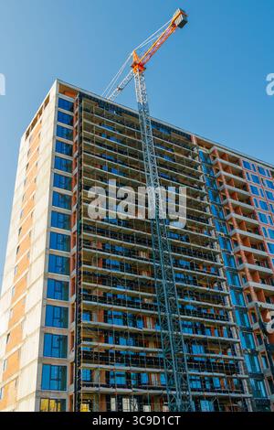 A tall residential building is under construction with a metal scaffold and a large tower crane, captured on a sunny day with blue sky. Stock Photo