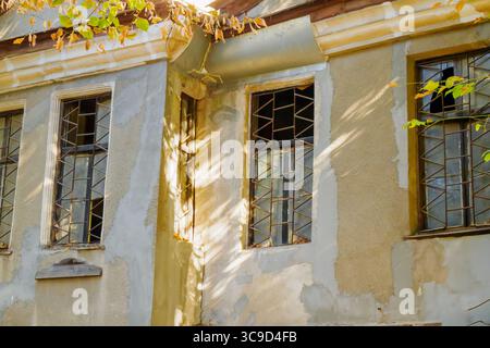 An aged urban building shows signs of wear with cracked plaster and barred windows, while soft sunlight filters through leaves onto the textured walls Stock Photo