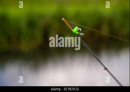 Close-up of fishing rod tip with bell bite alarm clipped on, blurred background of calm water surface Stock Photo