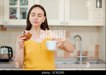 Peaceful moment as a young woman enjoys tea and a sweet bun in a warm, cozy kitchen, symbolizing comfort and slow living Stock Photo