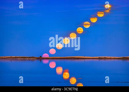 October 9, 2022, Alberta, Canada: The rising of the Full ''Hunter's Moon'' of October 9, 2022, with the Moon reflected in the calm waters this night at Crawling Lake Reservoir in southern Alberta. The Moon appeared very pink as it rose into the clear sky near the horizo, then turned more yellow as it rose into scattered bands of cloud. ..This serves as a goo demo of the Moon illusion and how the Moon does not change size as it rises â€” it is the same size on the horizon as it is higher up...This is a blend of 10 images taken 3 minutes apart with the RF70-200mm lens at 200mm and Canon R5, each Stock Photo