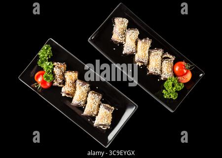 Eggplant rolls with cheese, garlic and sesame on black plates with herbs and tomato. Top view and isolated on black background. Stock Photo