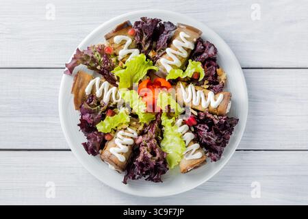 Eggplant rolls with cheese, garlic and white sauce and lettuce salad leaves on white plate and wooden table. Top view. Stock Photo