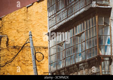 Textured urban study of Tineo, Spain: weathered iron and glass enclosed balcony with cracked panes set against a sunlit ochre wall and rustic wooden p Stock Photo