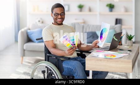 Black guy in wheelchair working as graphic designer online, holding color swatches palette, using laptop indoor Stock Photo
