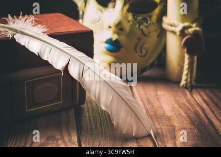 Literature concept. Feather on book near venetian mask and old scroll on wooden background. Stock Photo