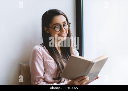 Leisure, education, favorite hobby concept. Cheerful woman reading book in cafe, sitting near window, free space Stock Photo