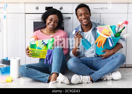 Smiling black couple sitting on floor, holding cleaning tools Stock Photo