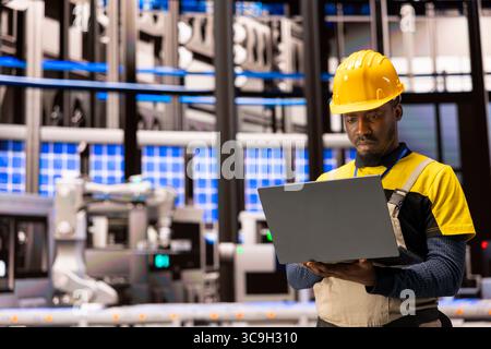 Black engineer monitors industry 4.0 system in industrial plant. Surrounded by robotic arms and conveyor belts, he uses laptop to ensure automated factory optimization, focusing on smart manufacturing Stock Photo