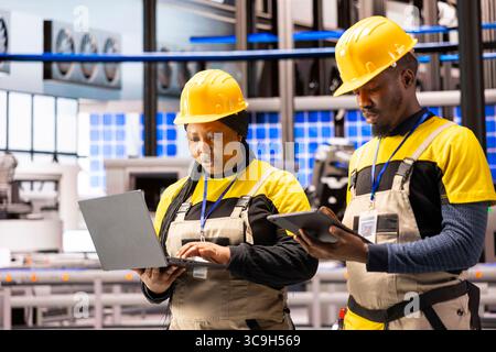 Industry 4.0 experts collect maintenance insights using laptop and tablet in advanced automated factory. Skilled male and female technicians overseeing optimization of smart manufacturing facility. Stock Photo