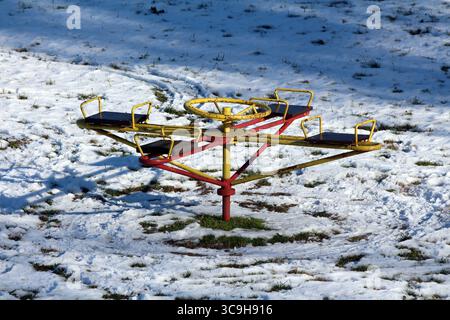 A yellow and red metal playground carousel stands on patchy snow and grass, its empty seats and shadows highlighting a cold, quiet winter day Stock Photo