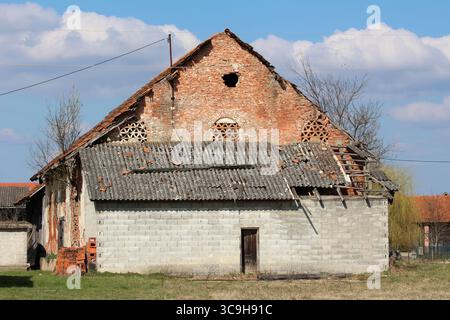 Old rural barn with exposed red brick, broken asbestos-cement roof, and patchwork repairs stands amid grass, revealing decades of weathering Stock Photo